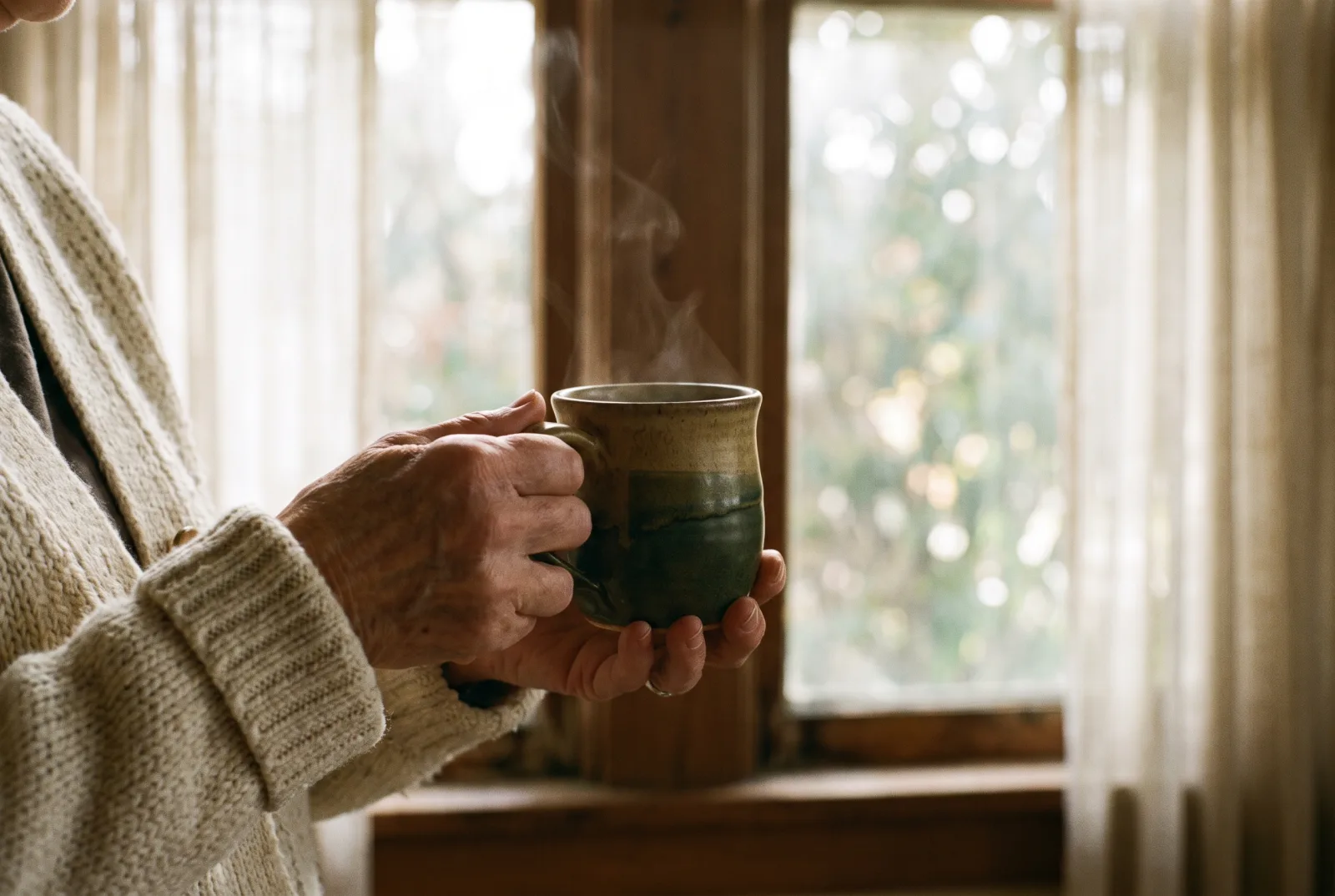 KI-generiertes Bild: Die Hände einer älteren Frau halten eine warme Keramiktasse, dahinter ein hohes Fenster mit weichem Licht.